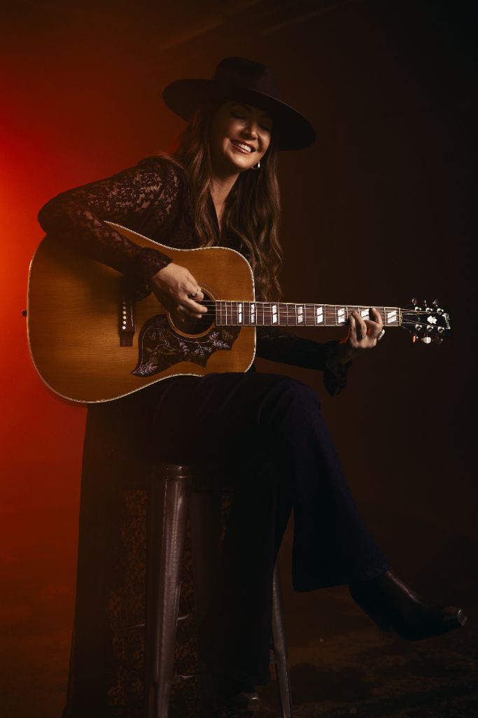 Kris Bradley seated on a stool, smiling, playing acoustic guitar, lit in warm red
