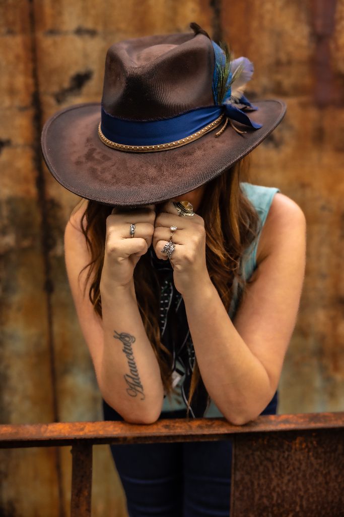 Kris Bradley, hat brim low, fists raised, head bowed, rings visible, tattoo on forearm, rusted wall behind her